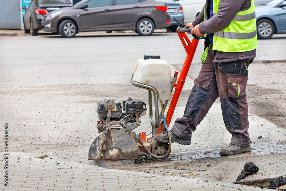 A worker cuts the old asphalt with a petrol cutter on the carriageway against the backdrop of a city street.