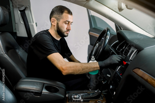 Close up portrait of professional car wash service worker in black clothes and protective gloves, cleaning car interior and control panel with special chemical anti dust liquid and soft brush © sofiko14