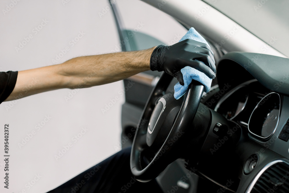 Cropped image of hand in protective glove of auto service worker, cleaning car interior, car steering wheel with blue microfiber cloth. Auto detailing and valeting concept.