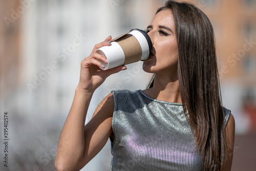 Caucasian girl in shiny gray top with a paper cup of coffee in hand. Brunette girl with dark hair drinking coffee on the city streets. Casual street fashion look.