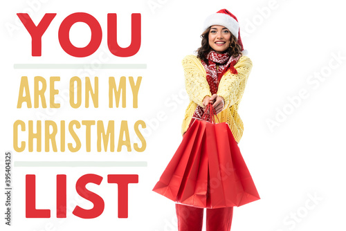  young woman in santa hat and scarf with ornament holding red shopping bags near you are on my christmas list lettering on white