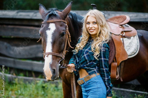 Smilng pretty young cowgirl. A cowboy style female in a plaid shirt standing next to the horse and holds the horse by the bridle.
