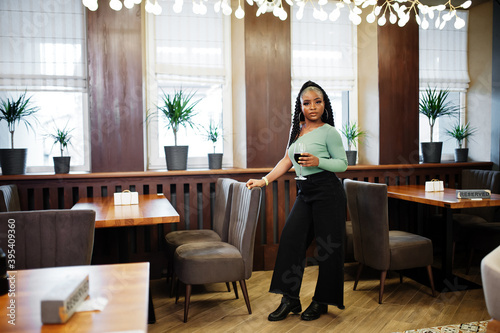 Portrait of attractive young african american woman wearing in green sweater and black jeans pose at restaurant with glass of red wine at hand. © AS Photo Family