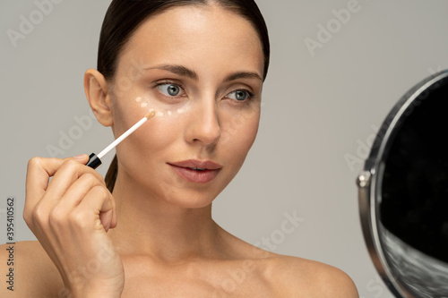 Woman with natural makeup applying corrector on flawless fresh skin, doing make up, looking at mirror. Girl after shower put concealer under eye area. Beauty face, skin care. Copy space, advertising. photo