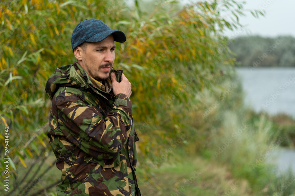 A man in a military jacket stands by the river bank. A man stands at the border.