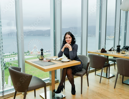 Elegant brunette woman sitting with coffee and notebook. Panoramic window with landscape. Business woman in cozy workplace. Woman taking notes in notepad.