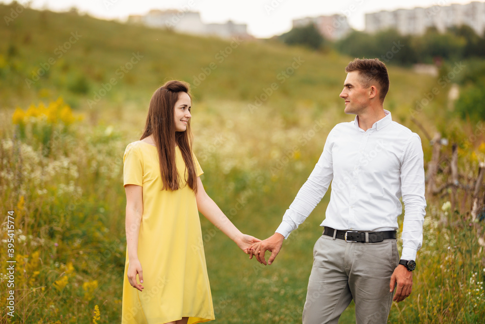young happy couple in love holding hands in a grass field in sunny day. girl in a yellow dress and a guy with a stylish haircut. portrait of couple walking on countryside young happy couple in love holding hands in a grass field in sunny day. girl in a yellow dress and a guy with a stylish haircut. portrait of couple walking on countryside