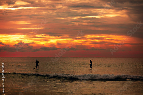 Fototapeta Naklejka Na Ścianę i Meble -  Sea sup surfing under amazing dark sunset sky. Two people on Stand Up Paddle Board. Orange sky. Paddleboarding Concept. Phuket. Thailand.