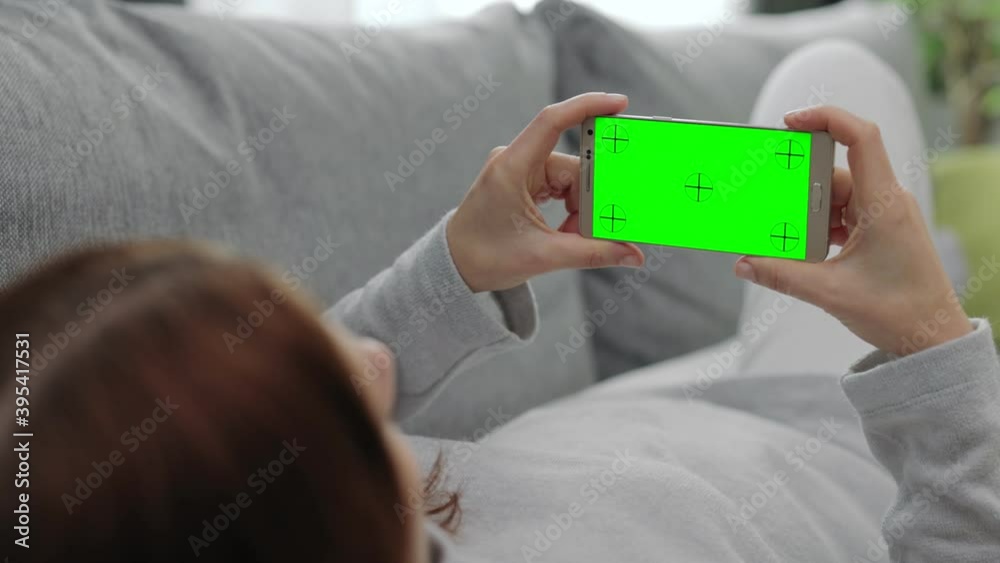 Back view of woman lying on comfortable couch and holding smartphone with horizontal green screen. Young brunette relaxing at home and using personal gadget. Back view of woman lying on comfortable couch and holding smartphone with horizontal green screen. Young brunette relaxing at home and using personal gadget.