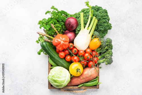 Variety of organic vegetables in brown box, broccoli cauliflower cabbage kale pak choy onions tomatoes. Healthy raw produce on white wooden table, top view, selective focus