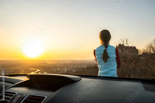 Young woman standing near her car enjoying warm sunset view. Girl traveler leaning on vehicle hood looking at evening horizon. © bilanol