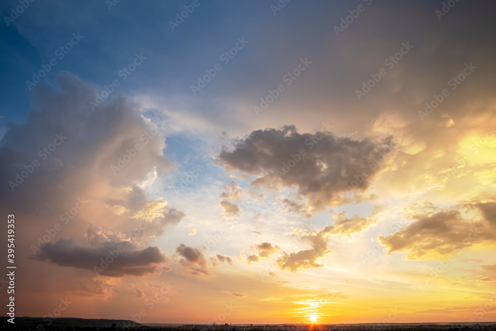 Dramatic sunset landscape of rural area with puffy clouds lit by orange setting sun and blue sky.