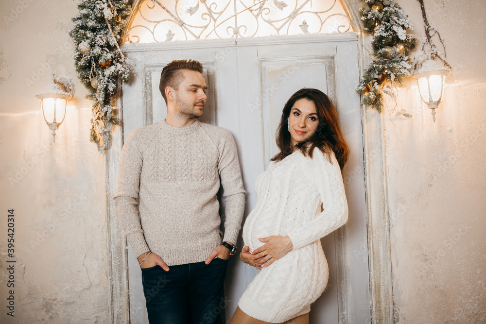 Portrait of happy couple, at the background Christmas decorations, charming husband with love look at the pregnant wife, winter holidays at home concept Portrait of happy couple, at the background Christmas decorations, charming husband with love look at the pregnant wife, winter holidays at home concept