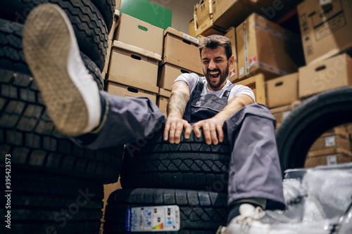 Smiling playful bearded tattooed worker sitting on tires in storage of import and export firm.