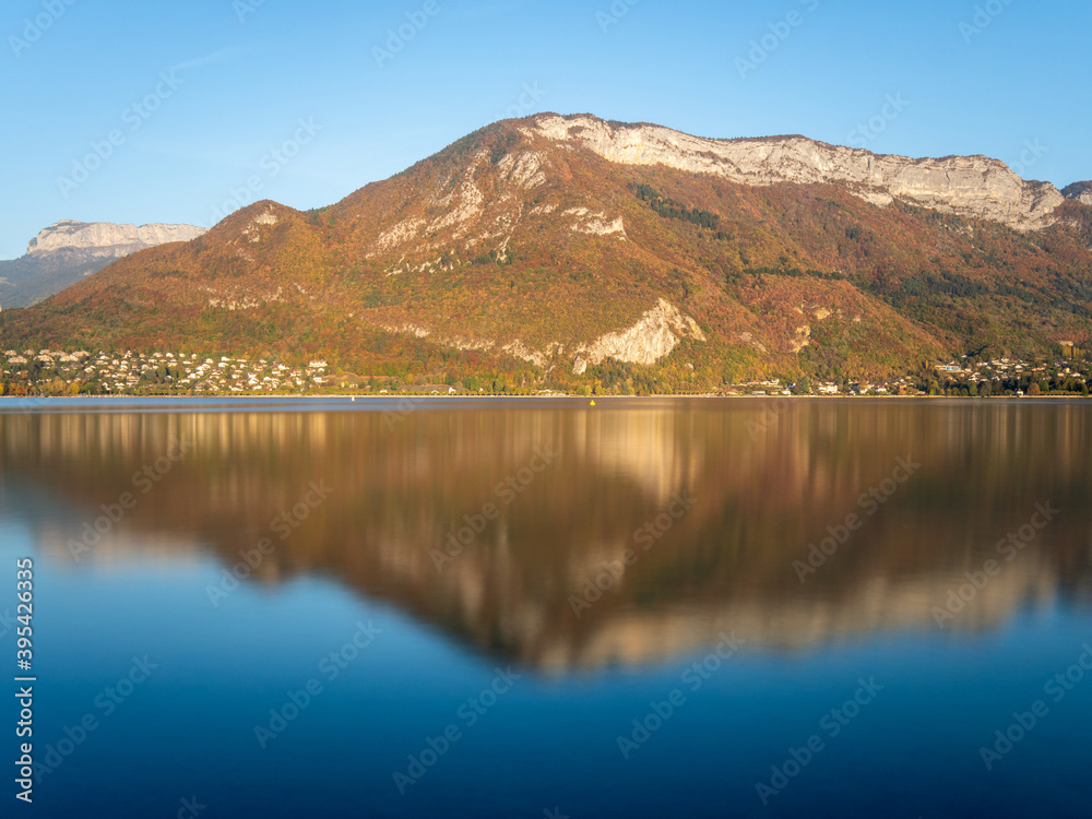 View of the mountains, behind Lake Annecy, in Savoie in the French Alps. It is autumn, the sky is blue and sunny. Reflection on the water. Landscape.