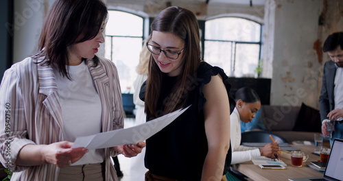 Beautiful middle aged Caucasian boss business woman helping younger coworker with papers at trendy loft coworking office