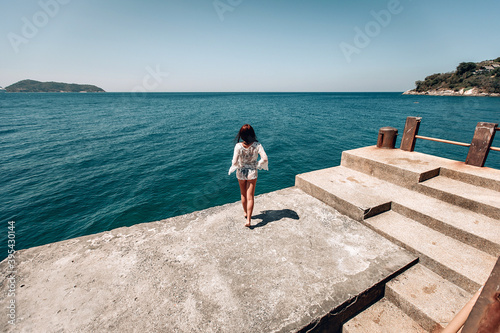 Slender girl in a short transparent white dress enjoying sun standing on wooden pier near the sea. Rear point of view. Phuket. Thailand