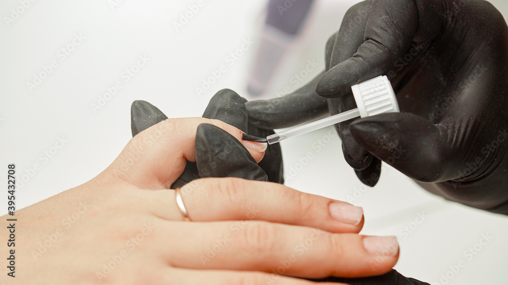 Fototapeta premium The manicurist applies a base coat to the nails. Close-up of the process of covering the nails with transparent varnish, manicure base, nail care, black gloves, cleanliness and care in the salon