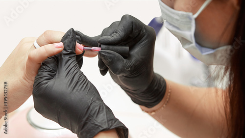 The manicurist applies a base coat to the nails. Close-up of the process of covering the nails with transparent varnish  manicure base  nail care  black gloves  cleanliness and care in the salon