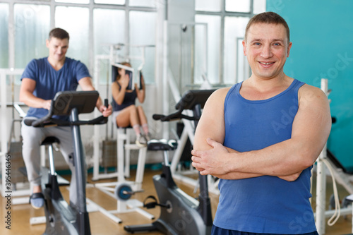 Portrait of cheerful sporty man posing at gym with his friends on background