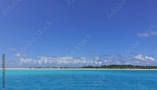 The calm ocean shimmers with aquamarine shades. An island with a sandy beach and palm trees is visible on the horizon. There are picturesque clouds in the azure sky. Maldives.