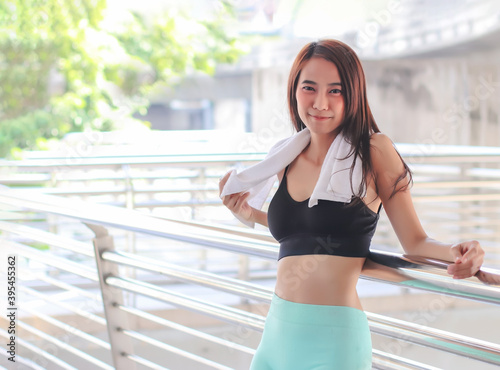 attractive Asian sportswoman in sportswear standing against bridge fence, smiling and looking at camera.