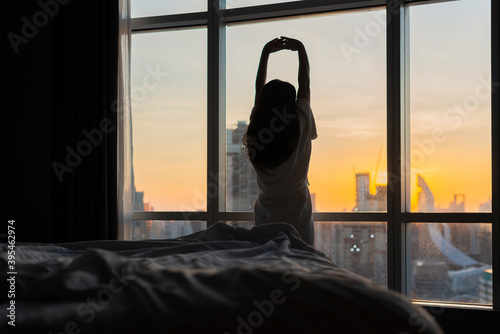 Asian woman standing and exercise near bed with beautiful sunlight in the morning and city scenery in the window.