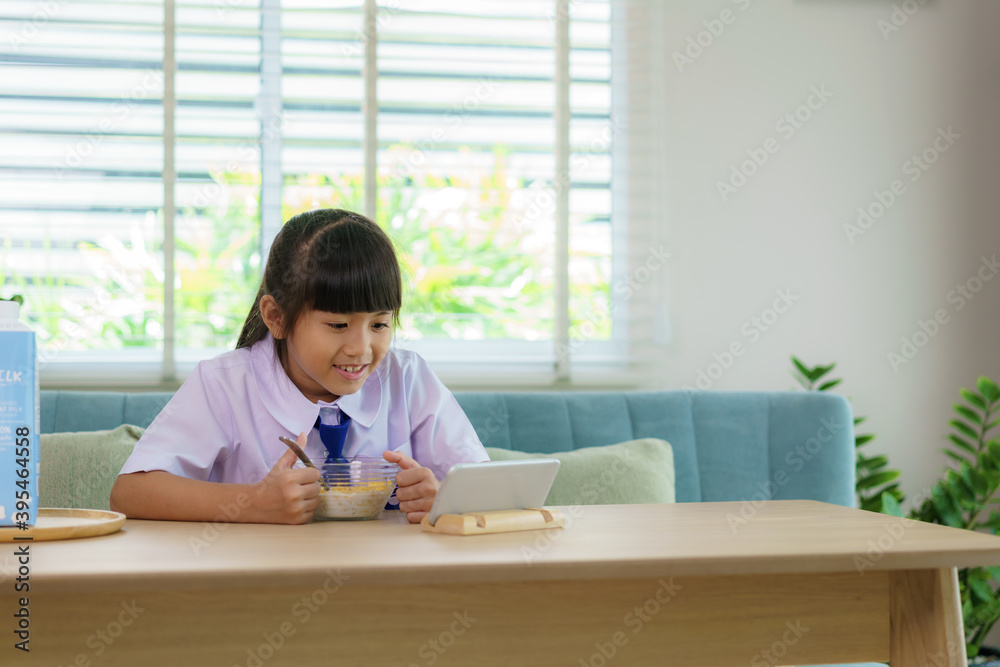 Asian elementary school student girl in uniform eating breakfast cereals with milk and looking cartoon in smartphone to distractions to eat it in morning school routine for day in life getting ready. Asian elementary school student girl in uniform eating breakfast cereals with milk and looking cartoon in smartphone to distractions to eat it in morning school routine for day in life getting ready.