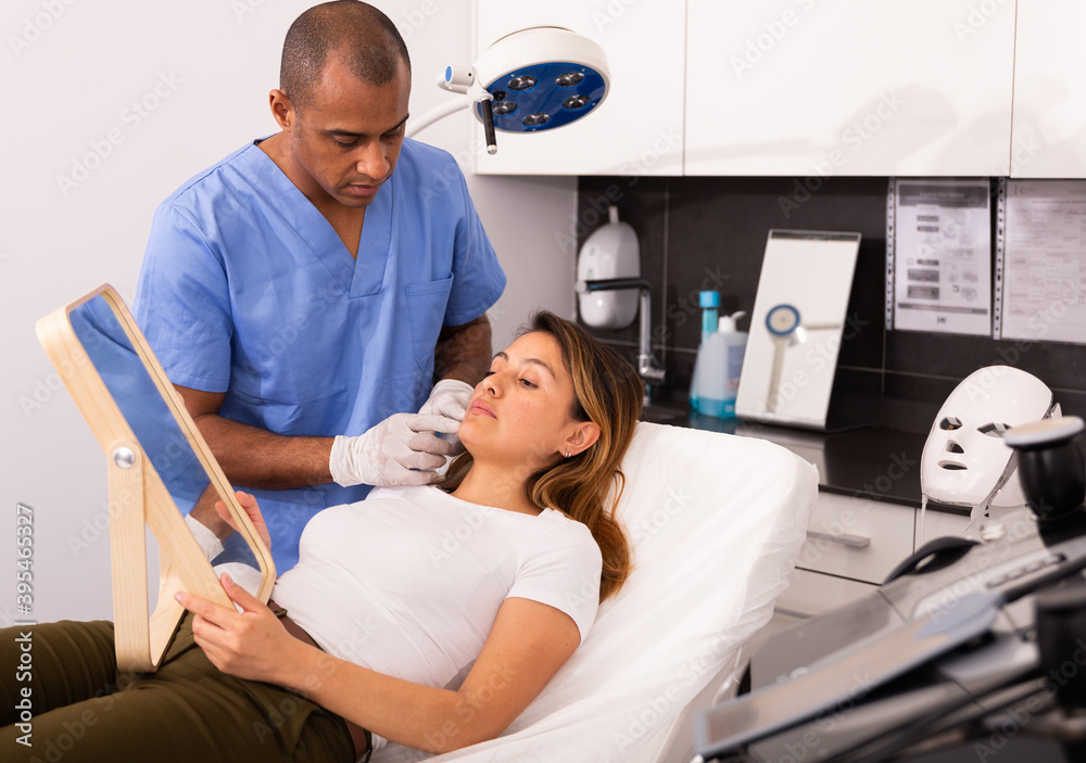 Obraz premium Focused Latina looking at mirror while qualified beautician explaining her future hardware facial procedure in medical esthetic office