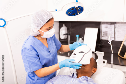Hispanic female beautician preparing male client for procedure of phototherapy, putting LED mask on mans face