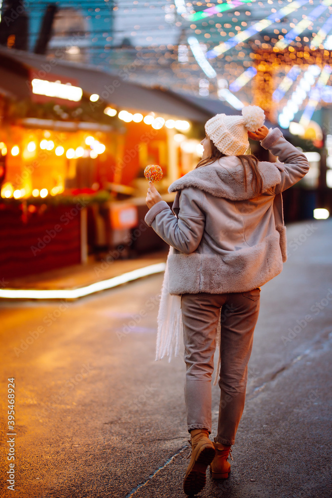 Young woman with caramel apple on Christmas market. Smiling woman in winter style clothes posing at festive street market. Lights around. Winter holiday.