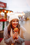 Young woman with caramel apple on Christmas market. Smiling woman in winter style clothes posing at festive street market. Lights around. Winter holiday.