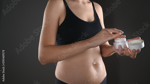 A young woman with loose abdominal skin and stretch marks holds a jar of cream in her hands. She is fat after pregnancy baby birth, studio isolated on grey background. Skin care concept.