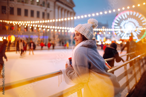 Smiling woman in winter style clothes with coffee near skating rink. Young woman enjoying winter holidays on Christmas market. Lights around.