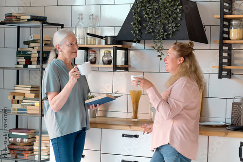 Friendly family viewing photos at home. Senior woman is enjoying a catch up with her daughter. They are drinking cups of tea in the kitchen. Elderly woman with female caregiver in living room photo