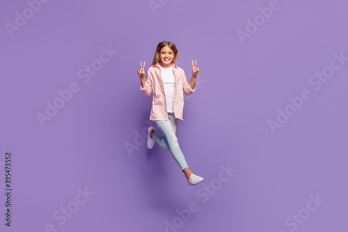 Full size photo of optimistic pretty girl jump show v-sign wear pink shirt jeans sneakers isolated on violet background
