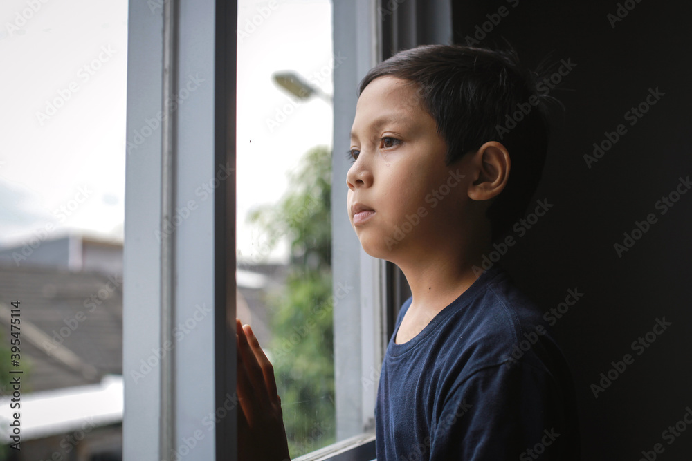 Stay at home coronavirus pandemic prevention. Sad asian boy standing on windowsill and looks out window. Stay at home social media campaign for coronavirus prevention
