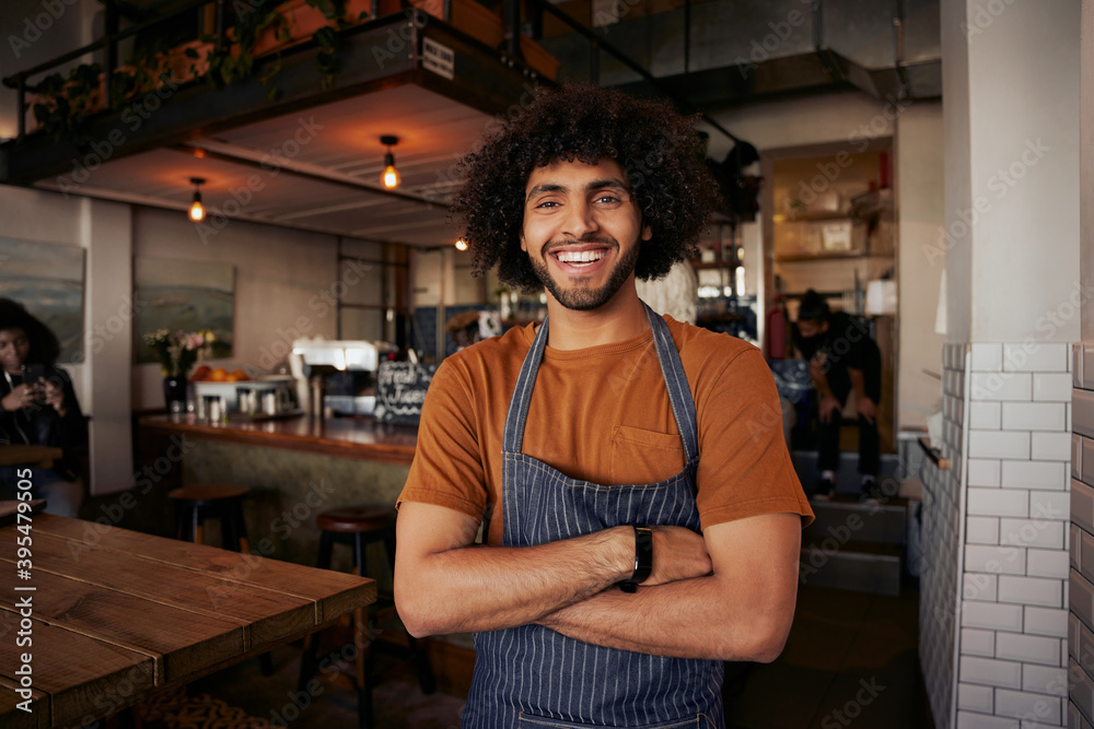 © StratfordProductions - Handsome young male waiter wearing apron standing with crossed arms in cafe looking at camera
