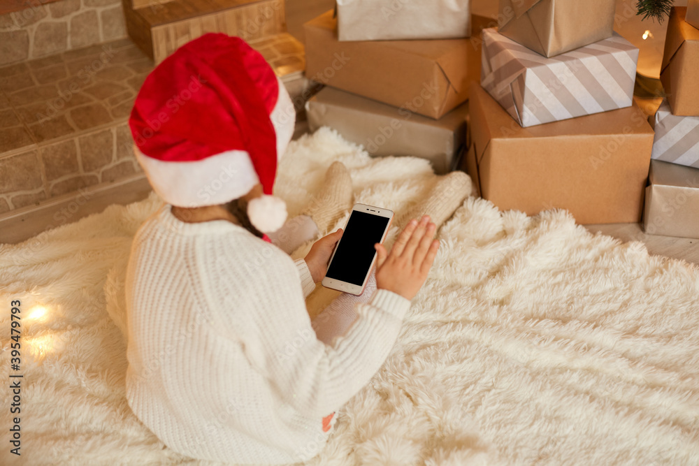 Obraz premium Little girl holding phone with blank screen while posing backwards, wearing white sweater and santa hat, sitting on floor on soft carpet. Merry Christmas and Happy Holidays!