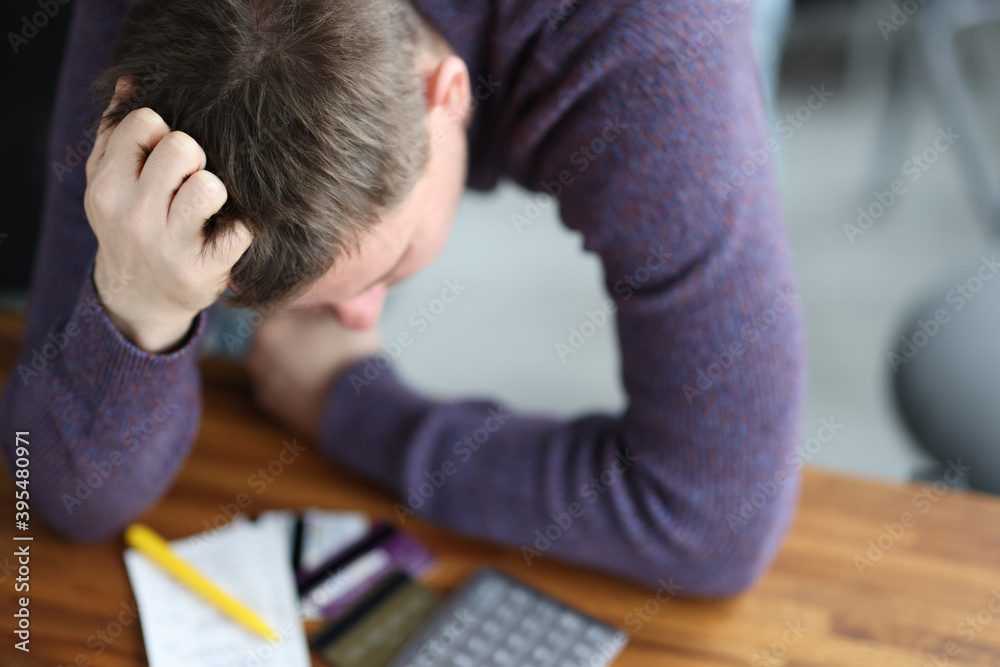 Sad man stands leaning over bank cards with calculator.