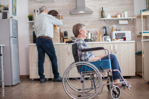 Disabled senior woman sitting in wheelchair in kitchen looking through window. Living with handicapped person. Husband helping wife with disability. Elderly couple with happy marriage.
