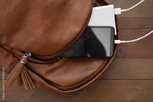 Charging mobile phone with power bank in backpack on wooden table, top view