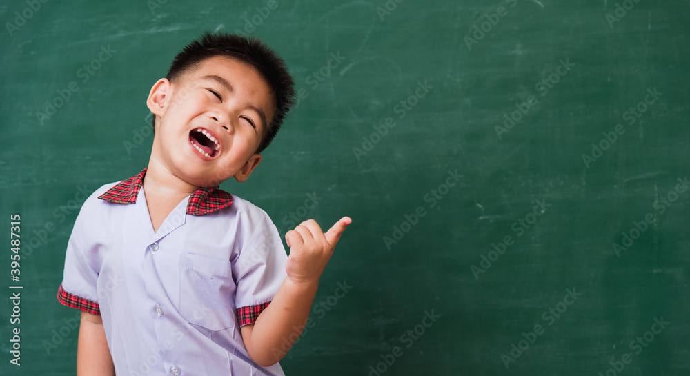 Back to School. Happy Asian funny cute little child boy from kindergarten in student uniform smiling point finger to side away space on green school blackboard, First time to school education concept Back to School. Happy Asian funny cute little child boy from kindergarten in student uniform smiling point finger to side away space on green school blackboard, First time to school education concept