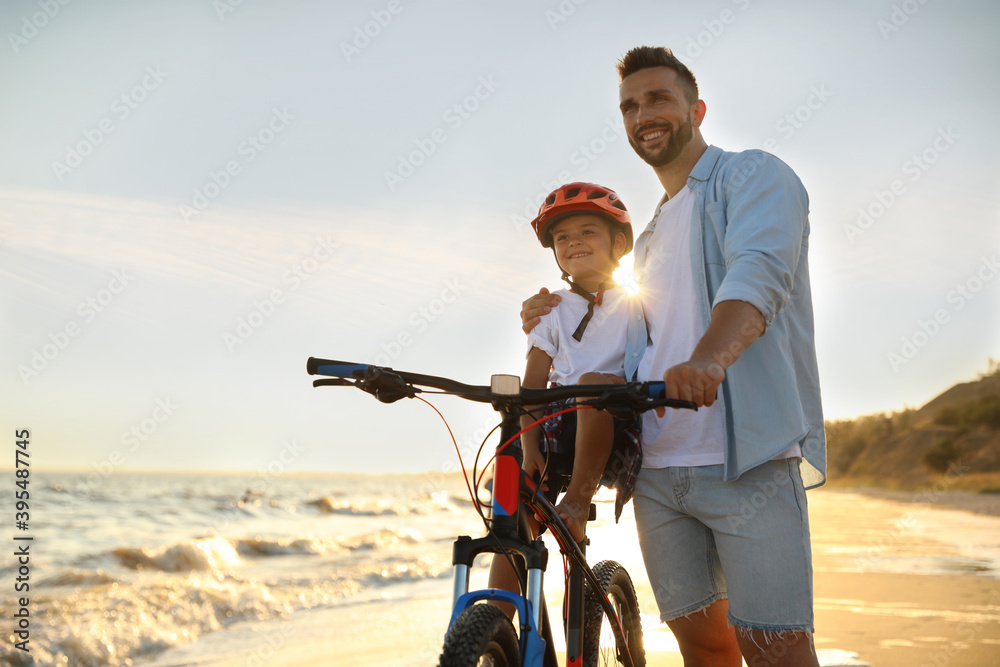 Fototapeta premium Happy father teaching son to ride bicycle on sandy beach near sea at sunset