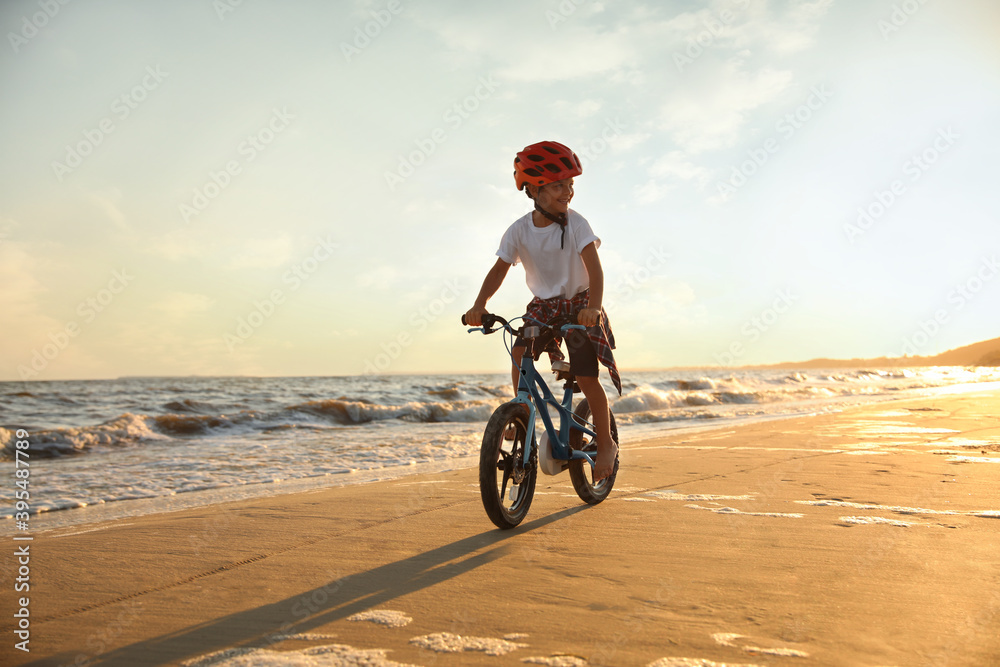 Fototapeta premium Happy little boy riding bicycle on sandy beach near sea at sunset