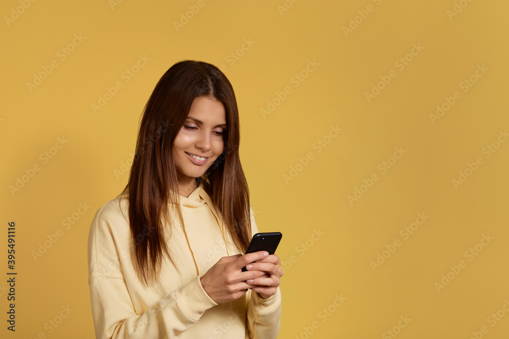 Attractive cheerful smiling caucasian woman in yellow hoodie holds mobile phone in hands, uses dating app, chats with boyfriend, looks joyful and interested, smiling, isolated on yellow background. Attractive cheerful smiling caucasian woman in yellow hoodie holds mobile phone in hands, uses dating app, chats with boyfriend, looks joyful and interested, smiling, isolated on yellow background.