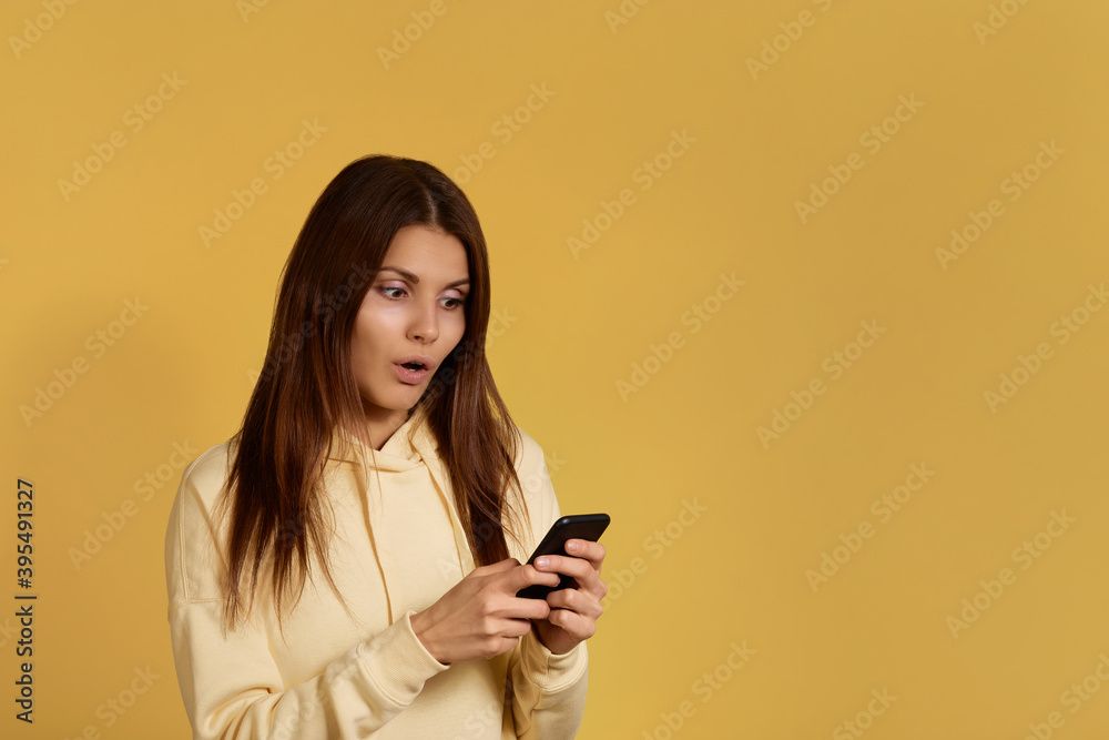 Attractive caucasian female in yellow hoodie with looks agitated at screen of mobile phone, impressed by message, stares at the screen, has surprised facial expression, isolated on yellow background. Attractive caucasian female in yellow hoodie with looks agitated at screen of mobile phone, impressed by message, stares at the screen, has surprised facial expression, isolated on yellow background.