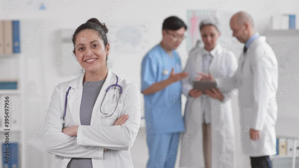 Waist up POV of young Mixed-Race female doctor standing in bright medical office, crossing her arms on chest, looking and smiling on camera. Blurred colleagues talking on background Waist up POV of young Mixed-Race female doctor standing in bright medical office, crossing her arms on chest, looking and smiling on camera. Blurred colleagues talking on background