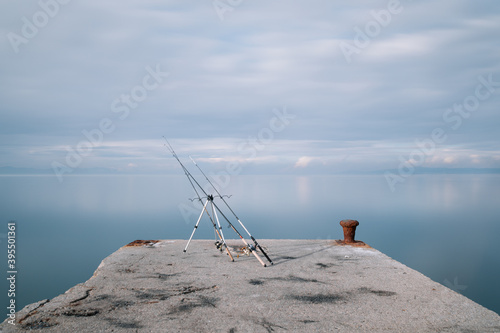view at the end of the pier overlooking the sea with a fishing rod line sat on a stand with nobody nearby near it sea is smooth cloudy skies in greece