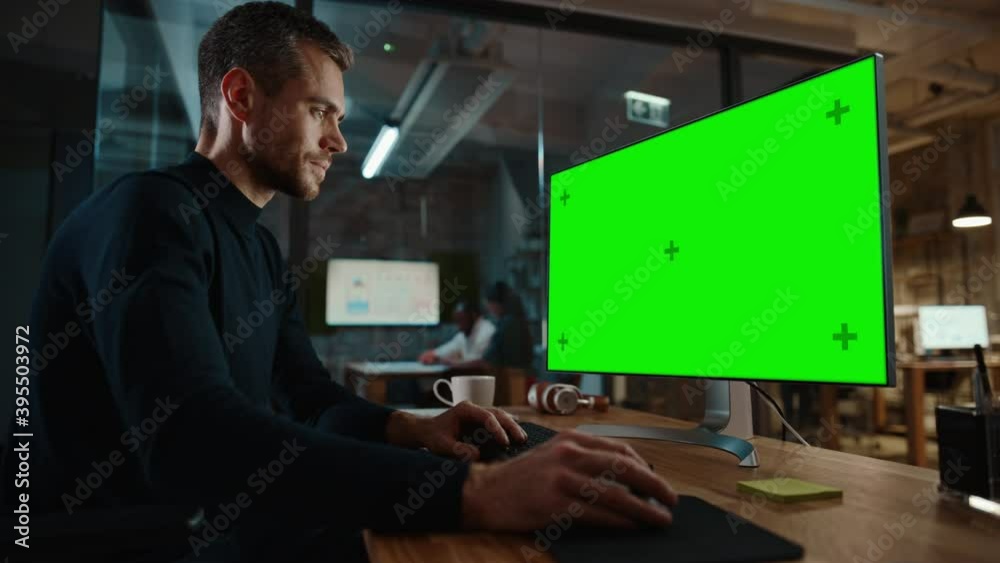 Young Handsome Specialist Working on Desktop Computer with Green Screen Mock Up Display in a Busy Creative Office with Colleagues. Male Manager with Trimmed Beard is Wearing a Casual Black Jumper.